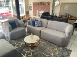 Light grey modern living-room showroom with a curved sofa, matching armchair, and blue patterned cushions; glass-topped coffee table on a geometric rug.