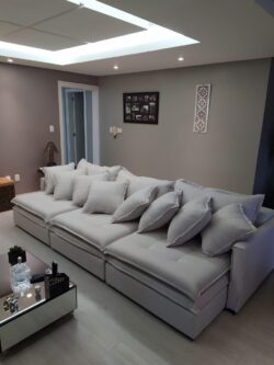 Large light-gray sectional sofa with multiple cushions in a contemporary living room, a glass coffee table with small decor, and neutral walls with wall art. A skylit ceiling provides bright overhead light.