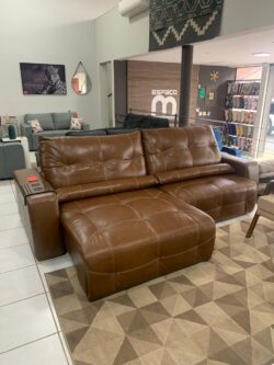 Brown leather L-shaped sectional sofa with tufted cushions in a furniture showroom, on a geometric rug with other seating and decor in the background.