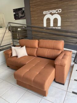 Brown fabric sectional with a chaise and two white throw pillows in a showroom setting, wood-paneled wall with 'espAÇO Motos' logo in the background.